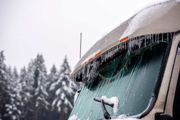 Ice-covered roof and windshield of big rig semi truck in snow and hanging icicles standing on parking lot with winter trees